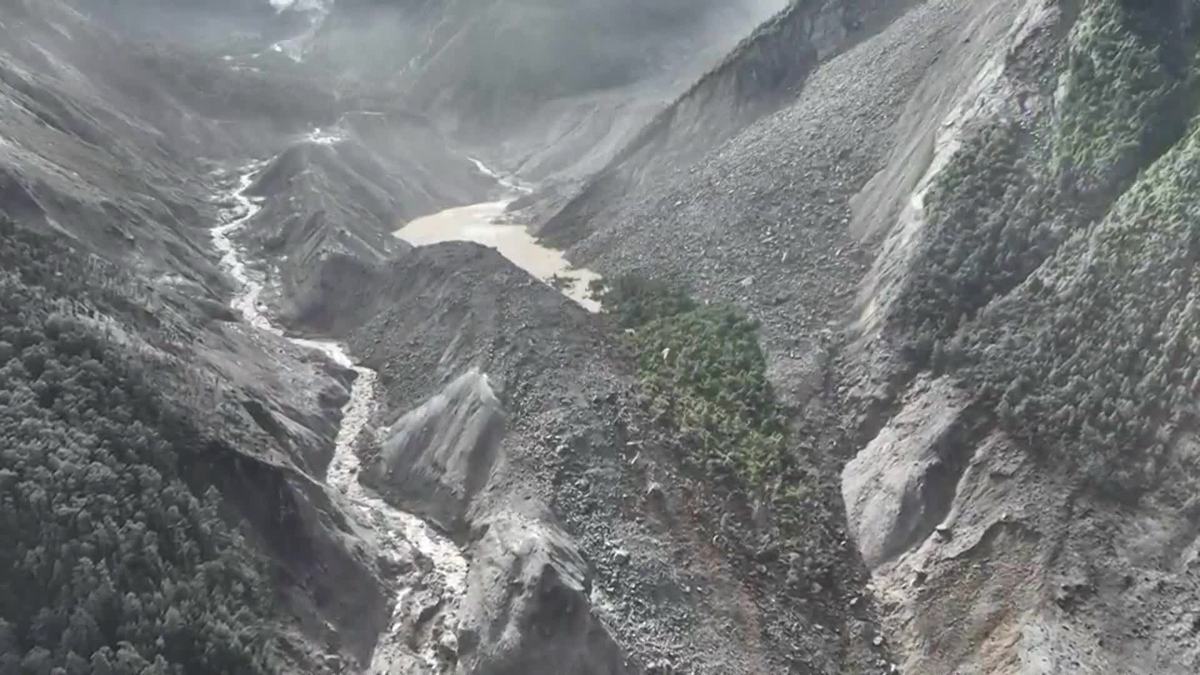 航拍：浑黄的堰塞湖水体被巨大的灰色冰岩崩堆积体拦在沟谷中，坝下有水流下泄，两岸山体被冲刷裸露