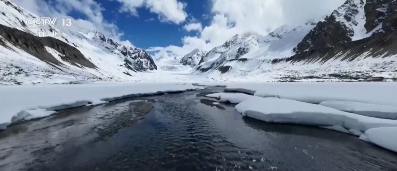 航拍冰川末端：远处是连绵雪山与冰川，近处冰面之上，融水汇成一条深色水道向前流去，两侧是残雪与裸露的冰面